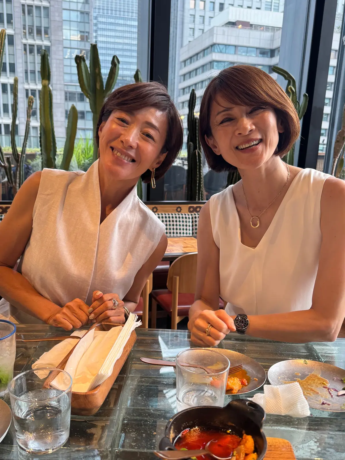 レストランで食事を楽しむ2人の女性。白い服を着て笑顔でこちらを見ている。テーブルには料理と飲み物が置かれ、背景に高層ビルと観葉植物が見える。