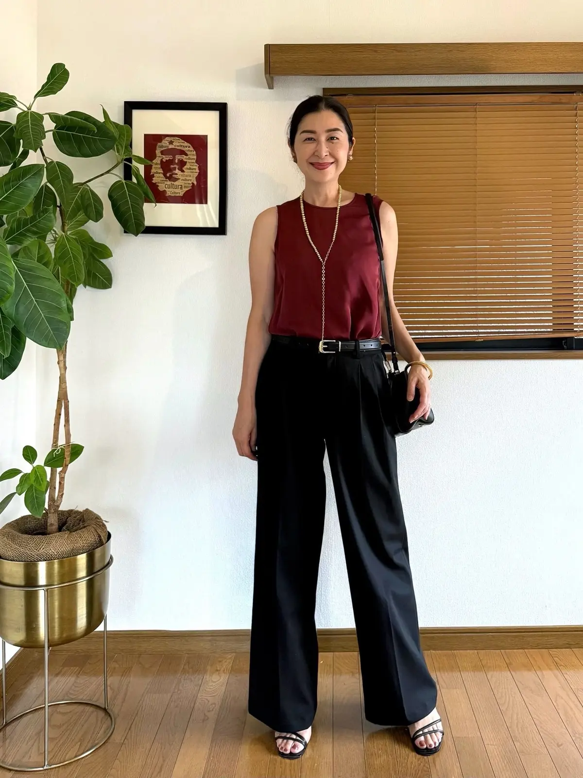 ユニクロスマートワイドストレートパンツ、赤いタンクトップに黒いパンツ、ベージュのベルトを着用した女性が立っている全身写真。右手に黒いハンドバッグを持ち、足元にはピンクのサンダル。背景には植物と窓が見える。