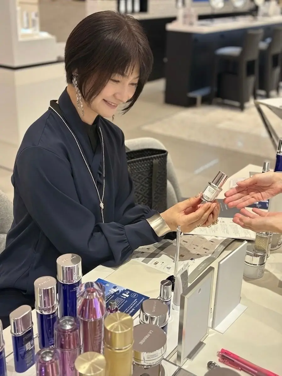 百貨店の化粧品カウンターで、女性店員が顧客に乳液を説明している様子。女性は黒いシャツを着ており、テーブルにはラ・プレリーの化粧品が並んでいる。