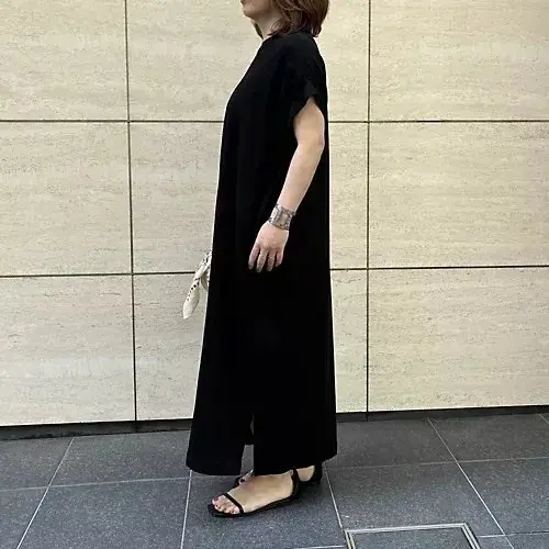 黒いマキシワンピースを着た女性が、左手で白い小袋を持ち、右手は壁に寄りかかり、ベージュのタイル壁の前で、黒いサンダルを履いている。