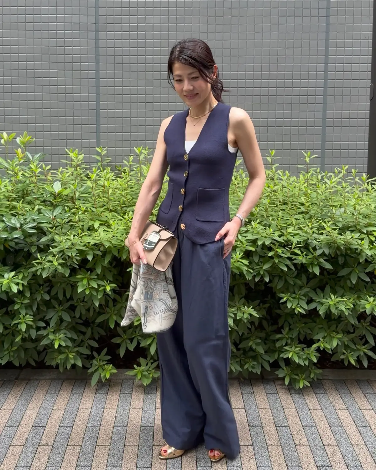 女性が青いベストとワイドパンツを着て、緑の低木の前に立っている。手にはベージュのハンドバッグを持っている。