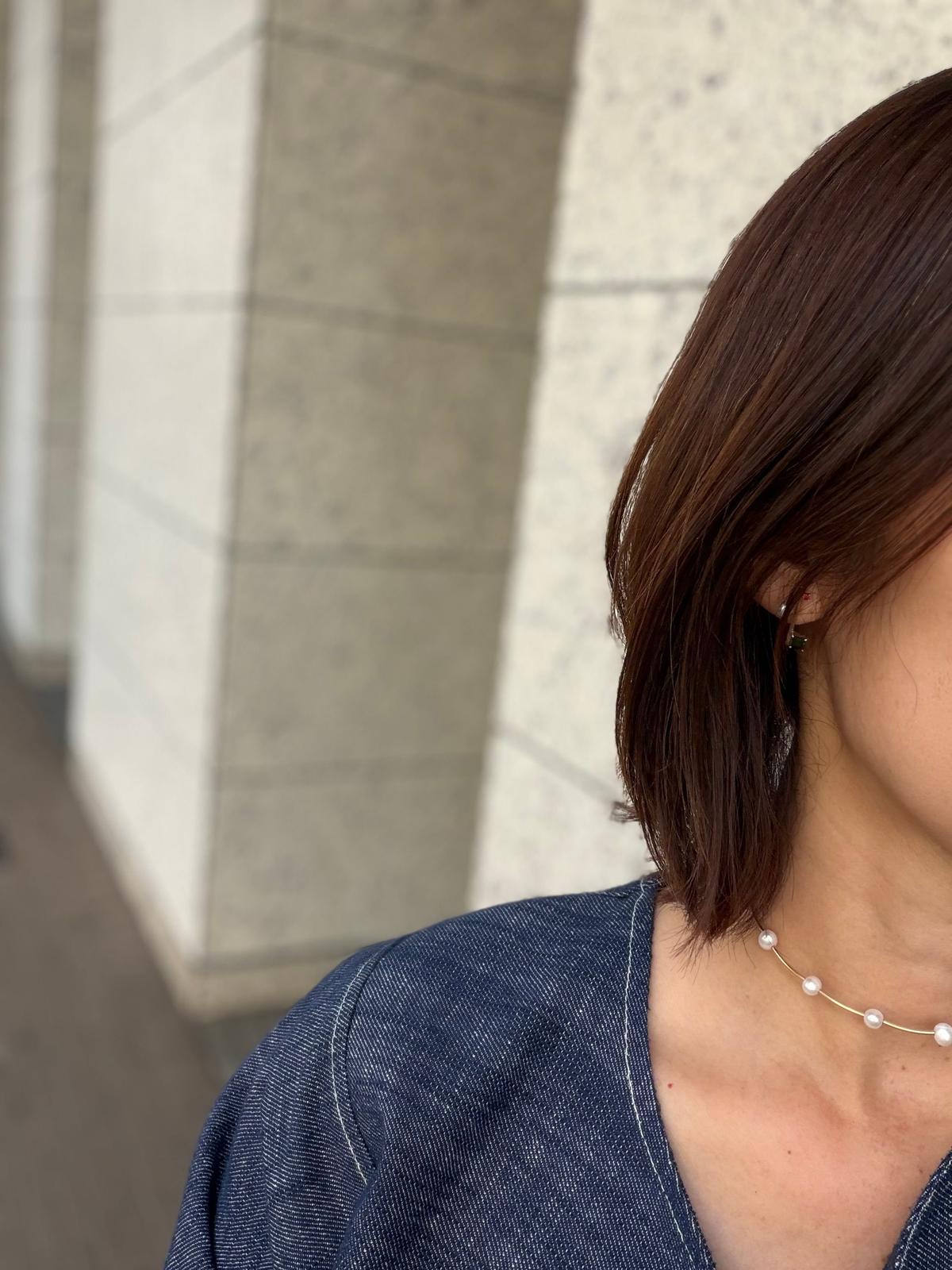 濃い茶色のボブヘアを肩まで伸ばした女性。デニムのシャツを着用し、首元にはパールネックレス、耳には控えめなピアスをしている。背景にはコンクリートと大理石の柱がある。