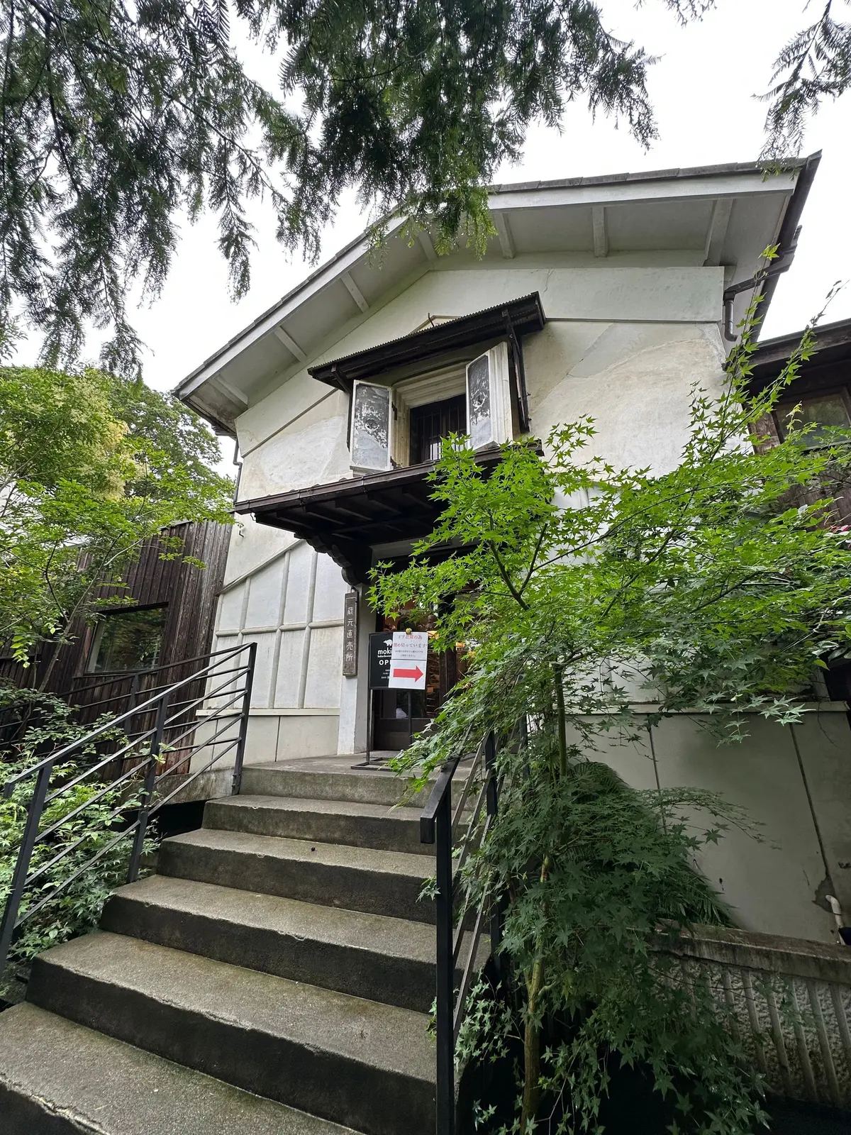 コンクリート階段と手すりがあり、木製のドアと看板が設置された白い建物。緑の木が建物周囲に生い茂っている。