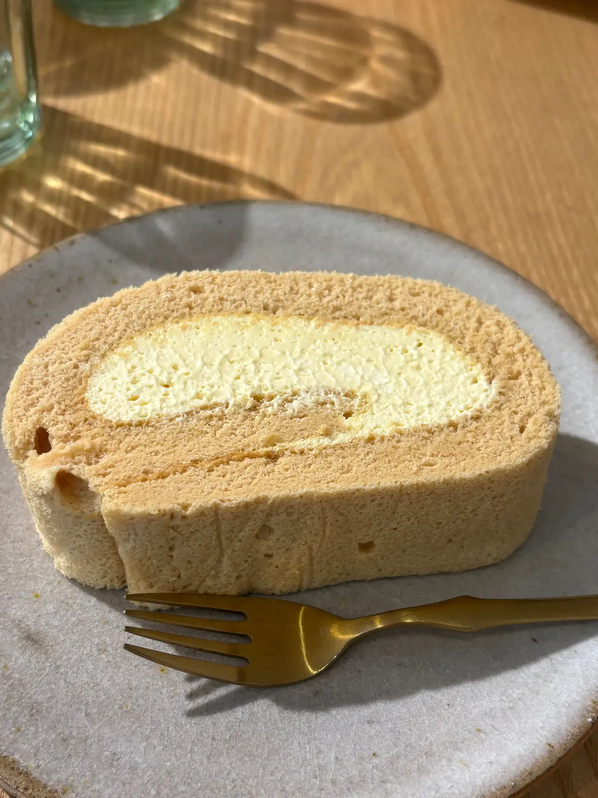 木目調のテーブルに置かれた、ロールケーキ。黄褐色のふわふわ生地で、淡い黄色のクリームが巻かれている。金色のフォークが添えられている。