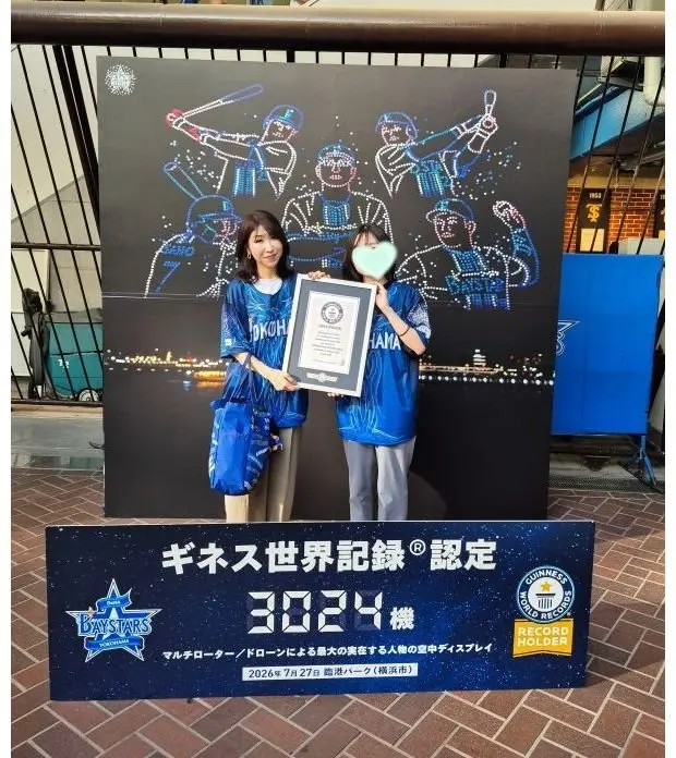 横浜ベイスターズのチームカラーである青色のジャージを着た2人の女性が、ギネス世界記録の認定証と記念プレートの前で笑顔を見せている。背景には、野球選手のイラストが描かれた壁がある。