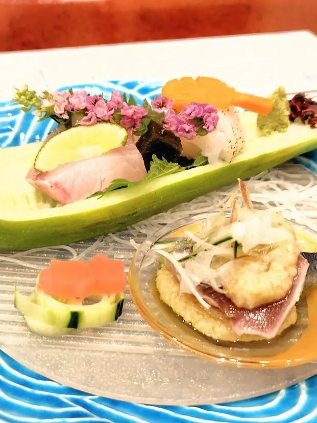 青い花模様のプレートに、キュウリにサンドウィッチした生ハムとライム、小さな花、野菜、寿司が並ぶ料理と、小さなボウルにカツレツと野菜の盛り合わせ