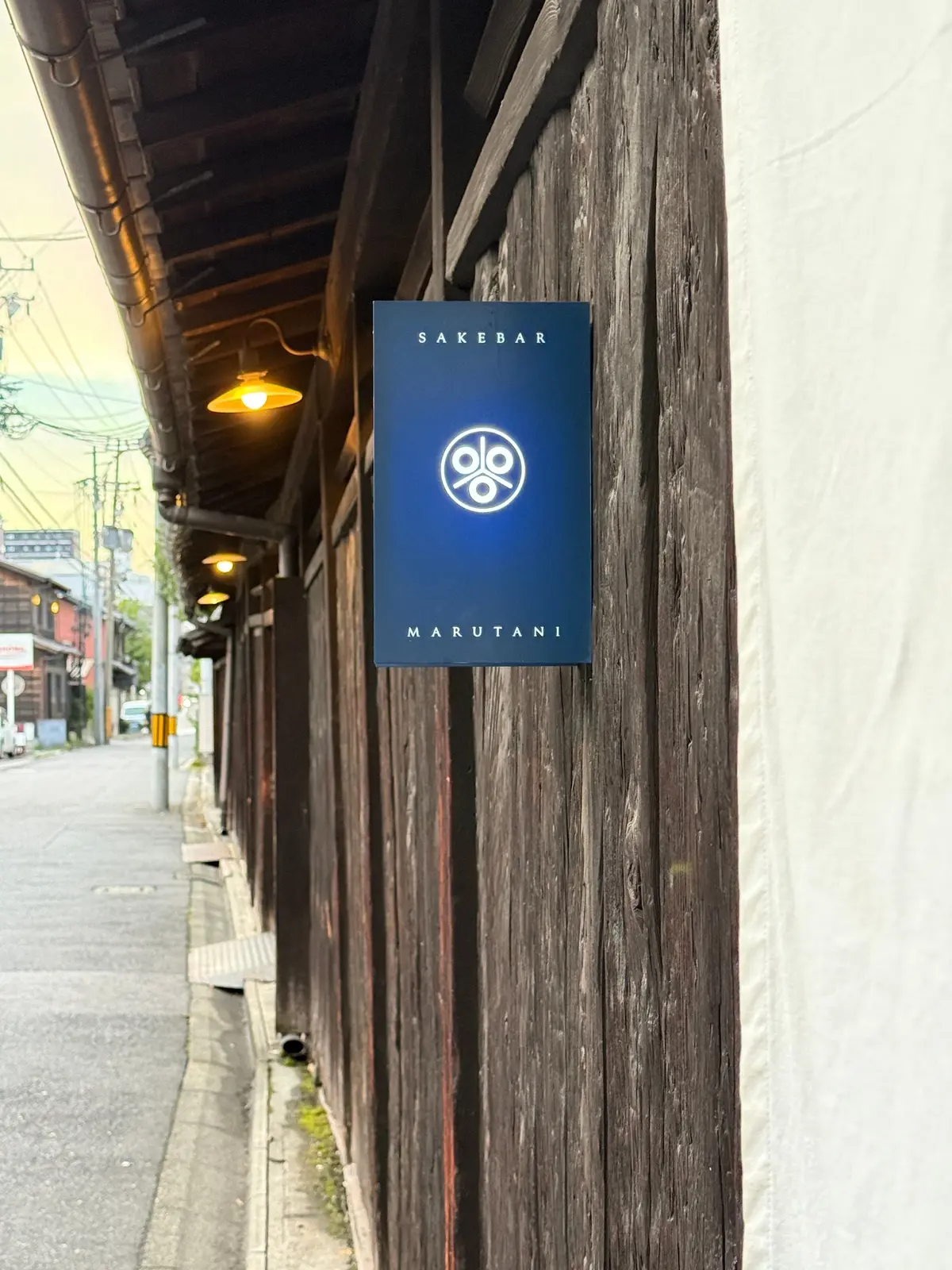 木造の壁に沿って、青色の「SAKE BAR MARUTANI」の看板が設置されている。看板は縦長で、中央に白地のロゴマークが配置されている。看板の左側には、電球型のランタンが数個吊るされている。背景には、細い路地と建物が見える。