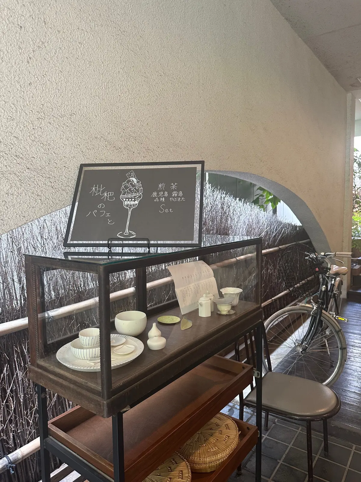 黒板のメニューとガラスケース、自転車が写る店内。黒板には「桃のパフェと前菜セット」の文字とイラスト。ガラスケースにはお皿に入った料理と飲み物が置かれ、右には自転車が置かれている。