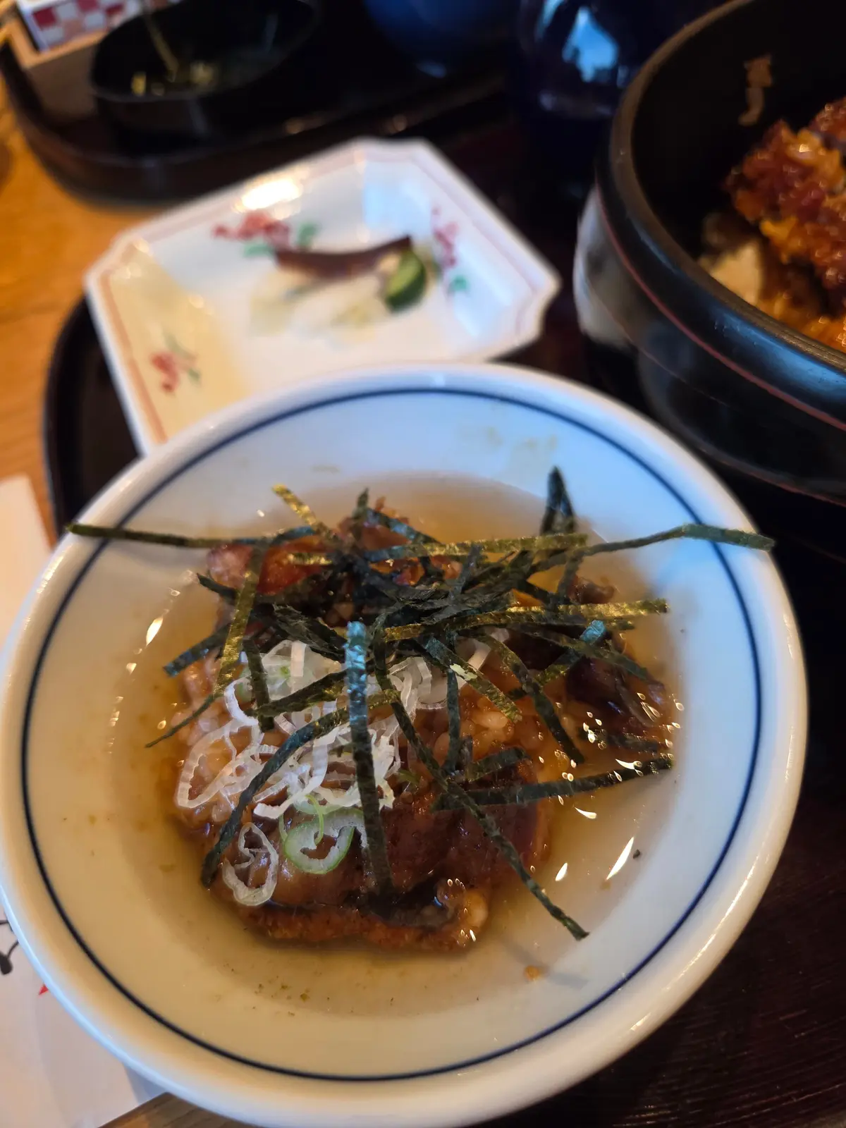白地に黒い縁取りの小皿に盛り付けられた、照り焼き風味の団子。上には細切りになったネギと海苔が散らされている。背景には、他の料理と食器が見える。