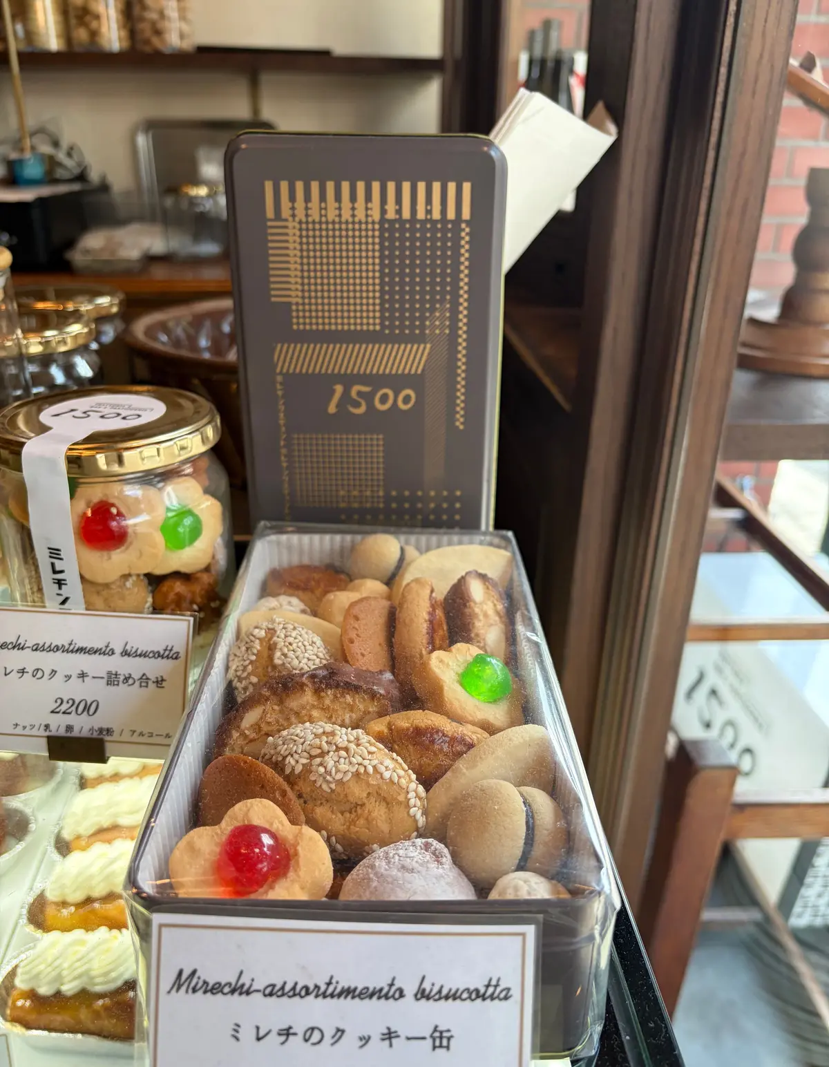 ガラスケースに並んだビスコッティの詰め合わせ。様々な形と色のビスコッティが並び、手書き風の価格表示カードが置かれている。隣にはジャムの瓶と、価格表示の黒い板も見られる。