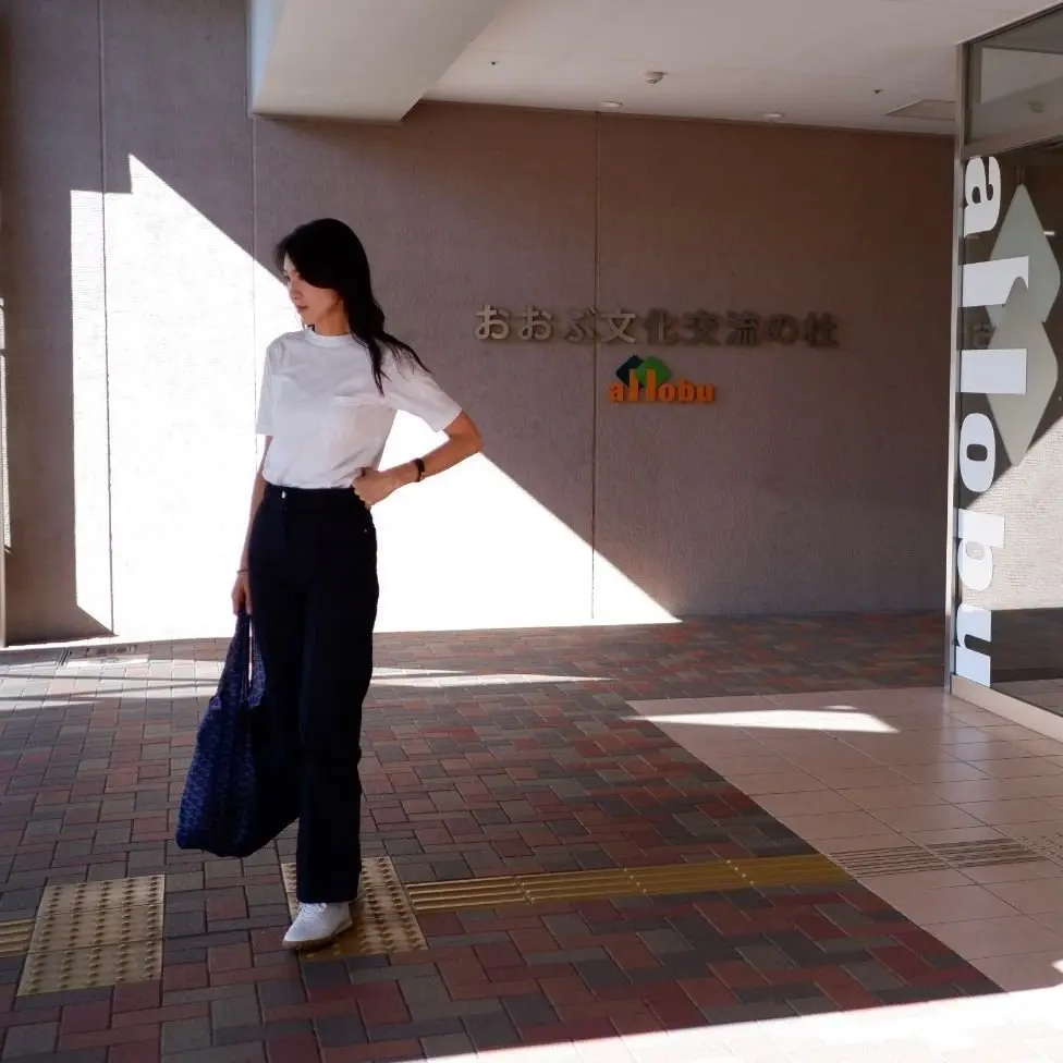 若い女性が、赤とグレーのタイル張りの通路を歩いている。背景にはガラス張りの建物があり、白いシャツと黒いズボンを着用した女性のシルエットが映っている。建物には「allobu」と書かれた看板がある。