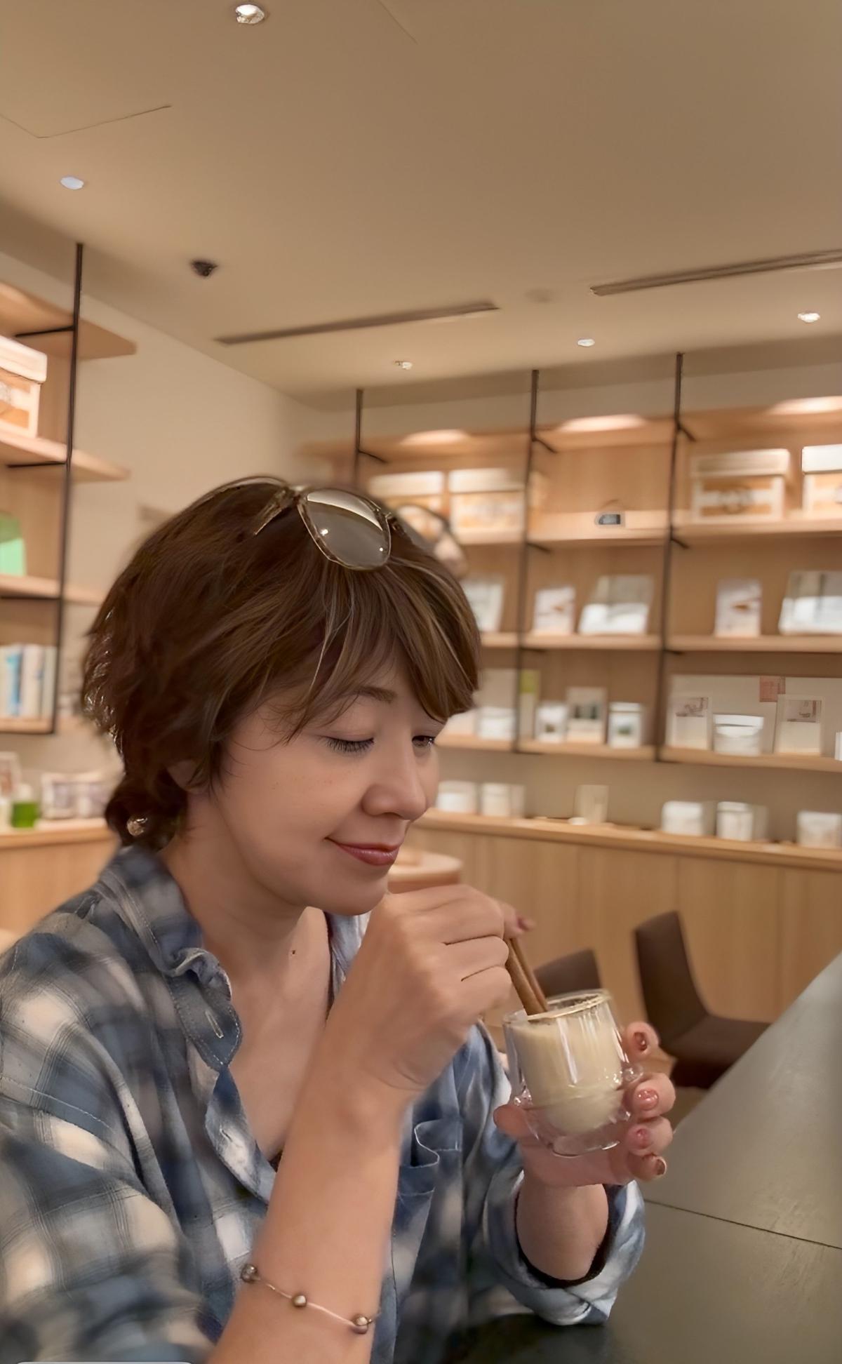 茶色のショートヘアの女性が、ガラスのカップに入った飲み物を楽しそうに飲んでいる。青と白のチェック柄のシャツを着ており、腕にはブレスレットを身につけている。背景には木製の棚が見える。
