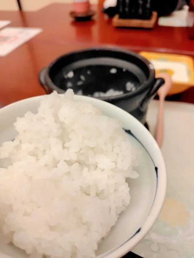 白いご飯が青い縁の鉢に盛り、黒い土鍋と木製の箸が背景にあるテーブル上